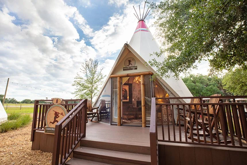 Luxe teepee at Westgate River Ranch Resort with a wooden deck, log railings and a rocking chair under a cloudy sky