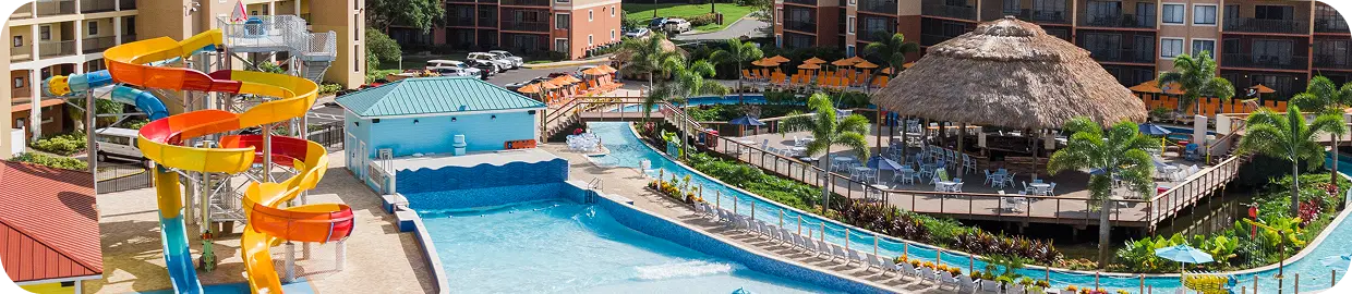 Resort pool area with colorful water slides, a winding pool and a thatched-roof pavilion beside hotel buildings