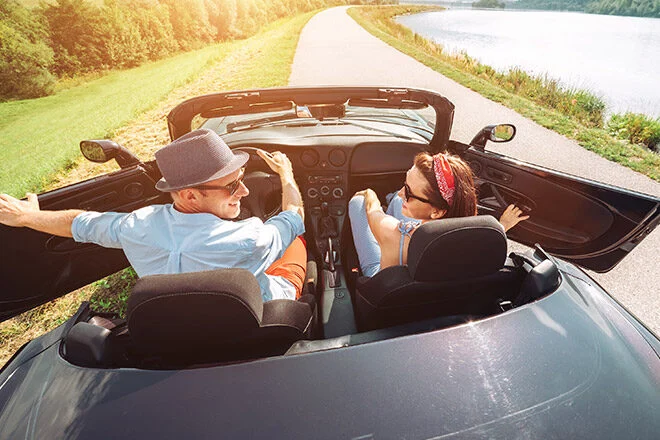 Couple in a silver convertible driving along a lakeside road with the man in a hat at the wheel