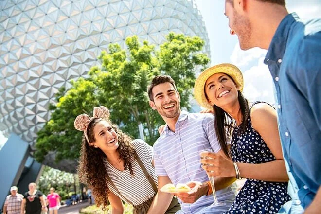 Friends laughing and sharing food in front of the silver geodesic sphere at Epcot, one wearing mouse ears