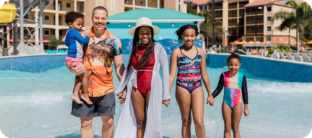Family wading in a resort pool with a child on dad's shoulders and resort buildings behind
