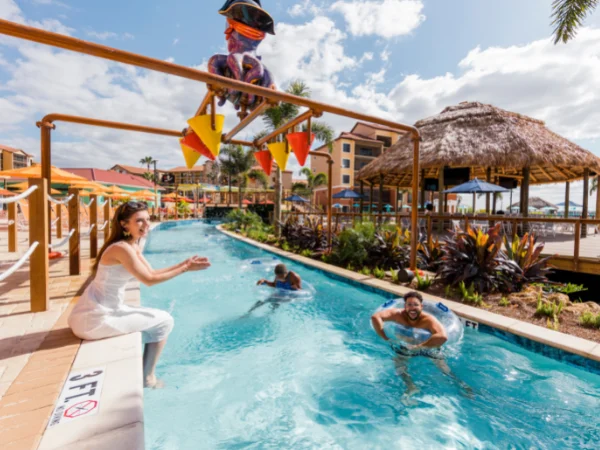 Woman sitting at the edge of a lazy river as a man floats by on a tube beside thatched tiki huts in Orlando