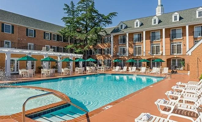 Outdoor pool with green umbrellas and white loungers beside colonial-style brick buildings in Williamsburg
