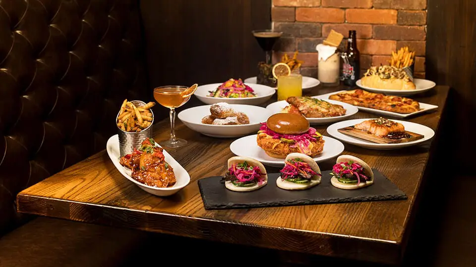 Spread of sliders, tacos, fries and flatbread with a cocktail on a wooden table in a dim restaurant