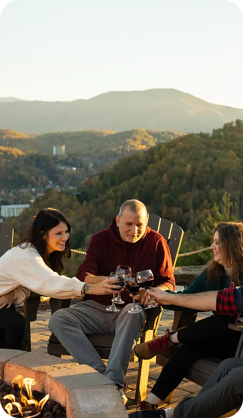 Friends toasting around a fire pit on a hillside with forested mountains behind at sunset